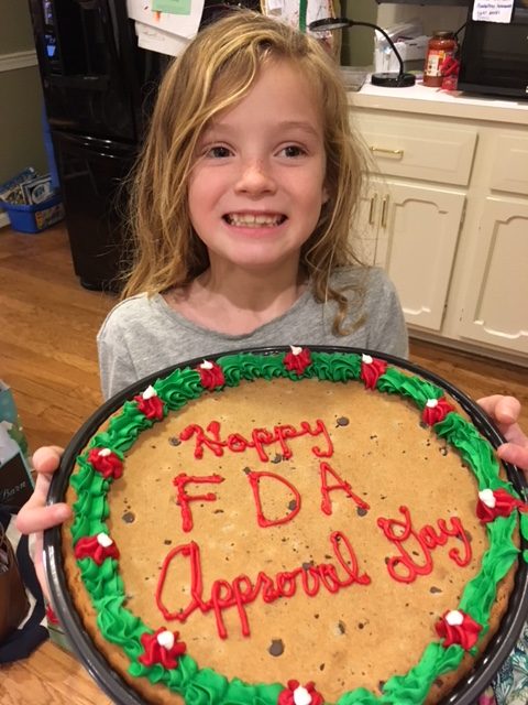Hannah Reif, 7, will be treated with LUXTURNA™ at Children’s Hospital of Philadelphia (CHOP) to cure her blindness caused by LCA-RPE65. Pictured: Smiling Hannah holding a cake that says Happy FDA Approval Day in red frosting.