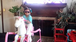 Young black child wearing cool sunglasses riding a rocking unicorn toy in front of a fireplace.
