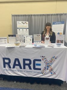 A woman stands behind an informational table at a RARE-X booth, with printed materials and signage displayed