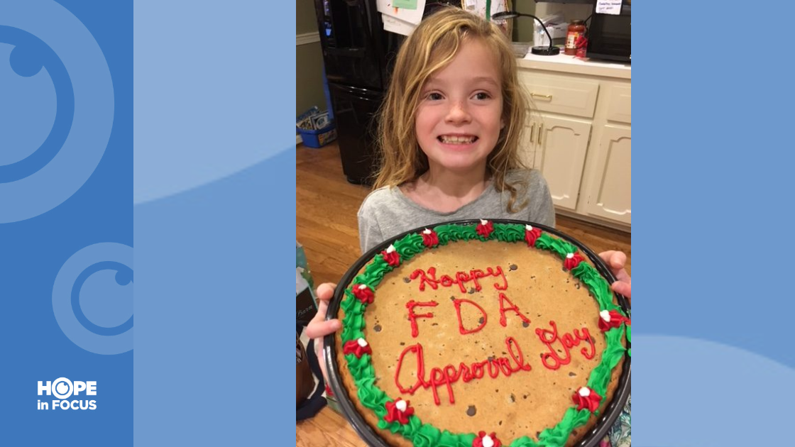 Smiling Hannah holding a cake that says Happy FDA Approval Day in red frosting.
