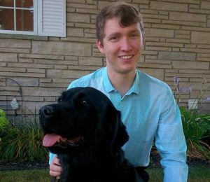 Jack McCormick in a blue shirt sitting next to his black lab, Jake