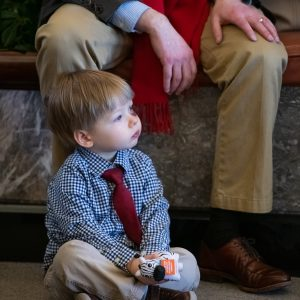 A little boy sitting cross legged in a blue plaid shirt and red tie, holding a NORD zebra stress toy.