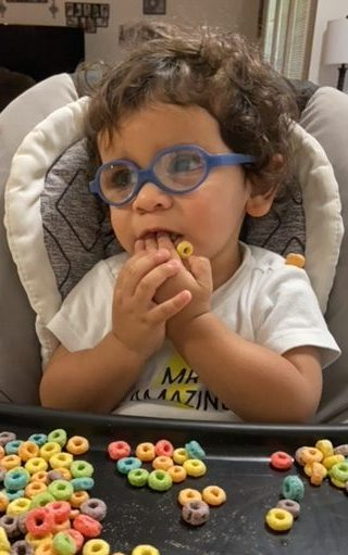 James with his hands in his mouth, sits in his high chair wearing a graphic tee.
