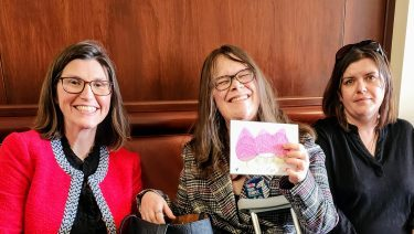 Three women smiling, Megan in the middle is holding up a drawing.