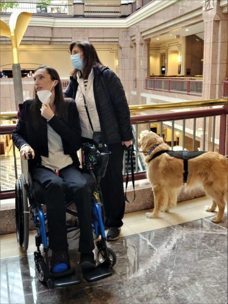 Kelly in her wheelchair, her mother is besides her holding her guide dog's leash.