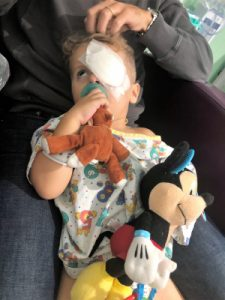 Jace in a hospital gown with a bandage on his left eye. He is holding his monkey binkie and Micky Mouse stuffed animal